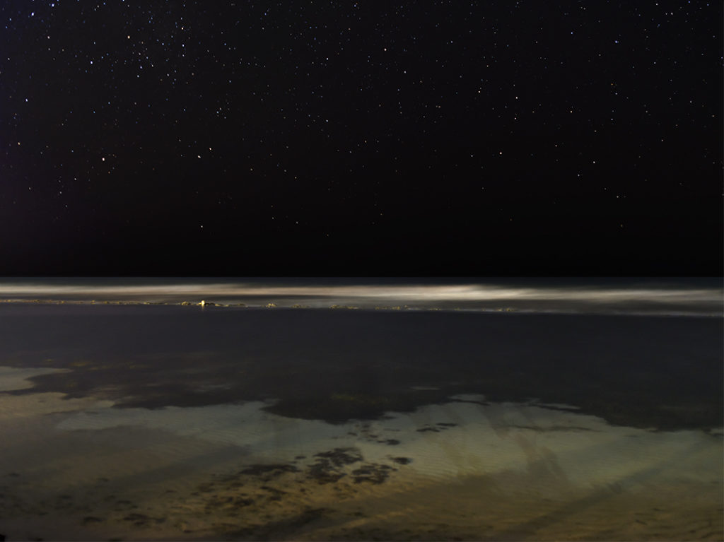 A Caribbean beach at night with a dark black sky, stars sparking and waters that are blurred and semi-transparent