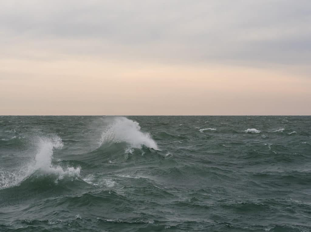 Waves crash on Lake Michigan. The water spraying into the air. The sky is peach in color, the waters a jade green.