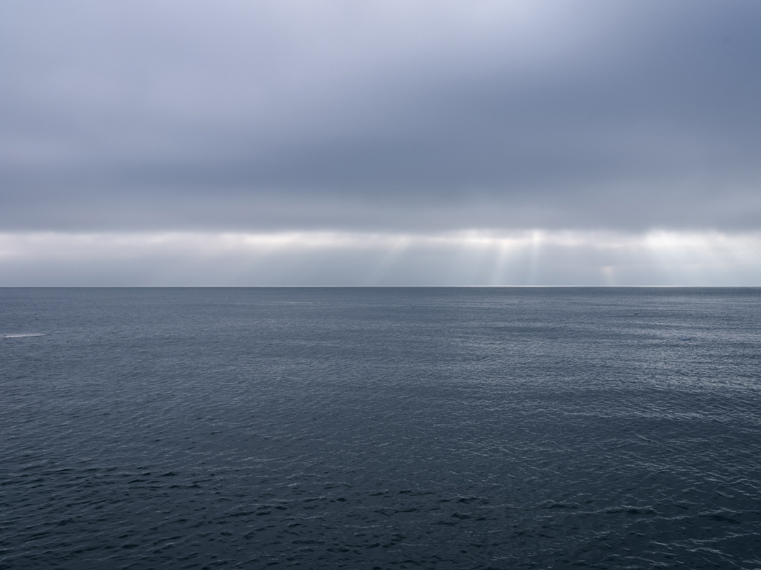 A slash of silver light cuts across at the horizon light far out over Lake Michigan, the light crisply bifurcates this photograph. A last piece of ice from recent storms floats out away from shore. This photograph is from January 29th, 2024.