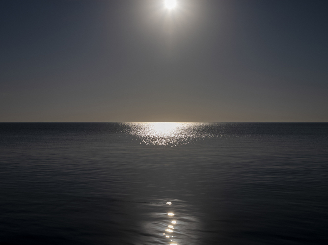 Near shore a series of glimmering spots of light, like stepping stones, mark a path out into the lake during a black lake dawn. Directing the eye to the horizon, where the sun creates a tightly glittering pool of light on the blue-black lake waters.
