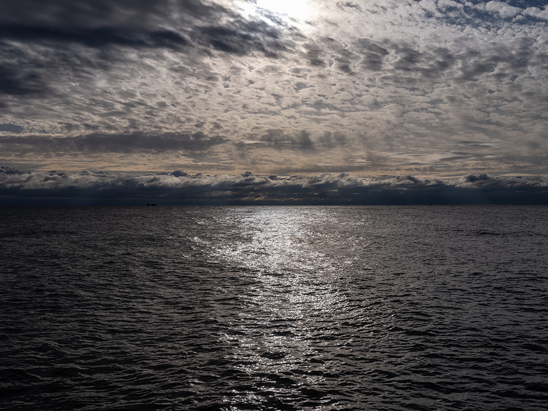 Lake and sky appear like chiffon rippled and bunched. Clouds and water give this photograph, Lake Series, October 20th, 2023, a heavily textured quality. Rendered in bronze, ochre and near-black tones. 