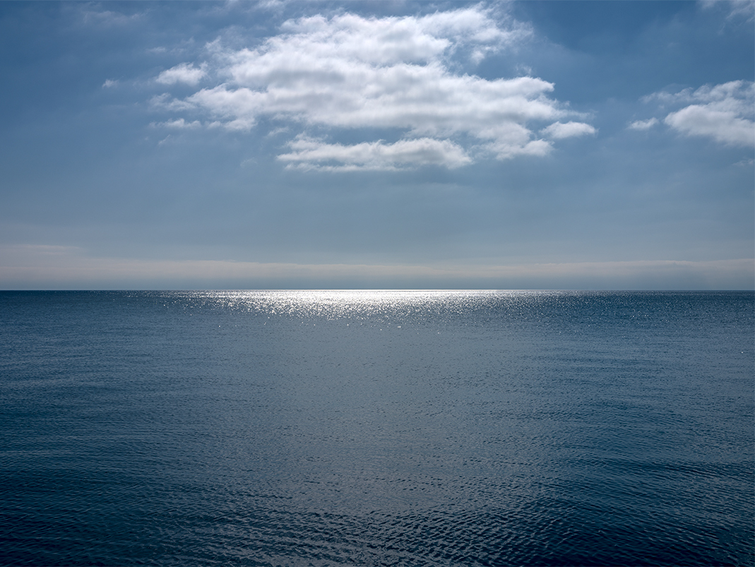 Clouds perch in the sky over Lake Michigan. The sun sears a white hot horizontal slash across the surface of the water near the horizon. This photograph is from the Lake Series, April 21st, 2022.