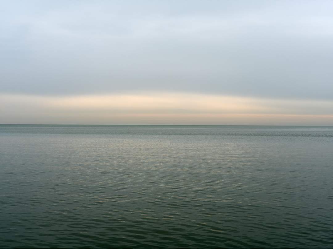 Dawn blooms on the lake, as the sky is beginning to come into light as I photograph in spring of 2020. As the day progresses the landscape will transform into deep blues, the softness of the sky seen here disappearing by mid-morning. But for now the lake and sky are rendered in peach and warm faded ochres alongside shimmering deep greens.