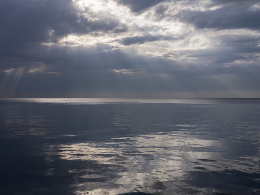 Still water, the lake is almost unmoving. Conditions that help create an abstract painterly quality to Lake Michigan’s water. The overhead light from the sun is filtering through clouds gently separating in the sky. Subtle shifts from dark to light and back again, happen throughout this photograph. An ethereal morning on Lake Michigan.