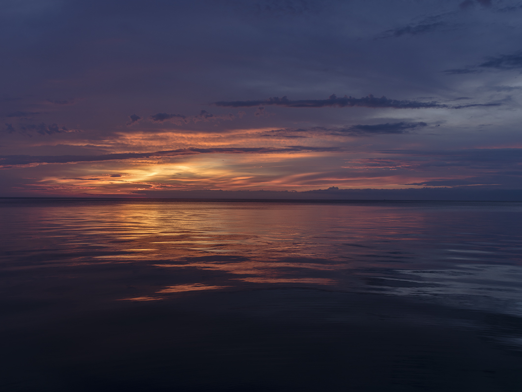 The sky breaks open over the lake and the sun at dawn is slashing through the clouds. Spilling its light in rich warm colors. Velvet tones of gold and red spread across the waters of Lake Michigan in this photograph from the Lake Series.