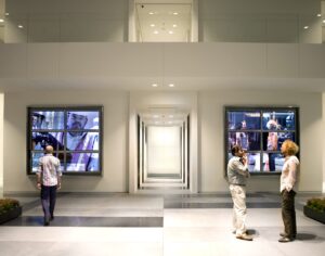 From Here, installed at One Arts Plaza by artist Lincoln Schatz, features two large video walls flanking an elevator core in a white lobby space, the lobby is shown on the screens