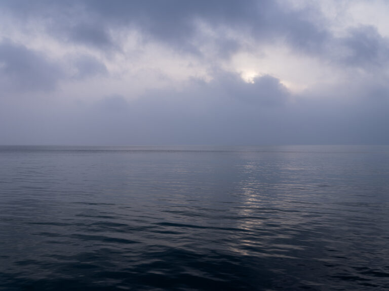 Purple hued Lake Michigan on an overcast morning where the clouds are just masking the sun behind them in a photograph from the Lake Series by Lincoln Schatz.
