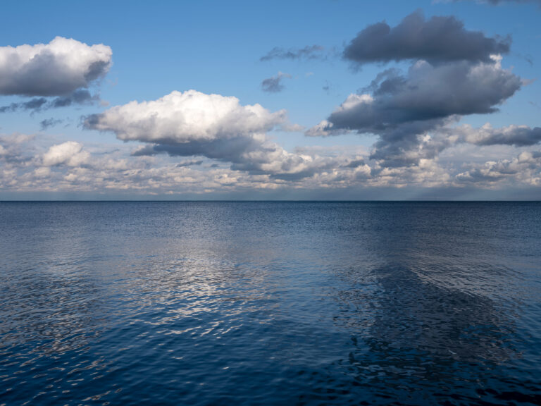 Clouds perch in a blue sky over a still Lake Michigan in a photo from the Lake Series by Lincoln Schatz.