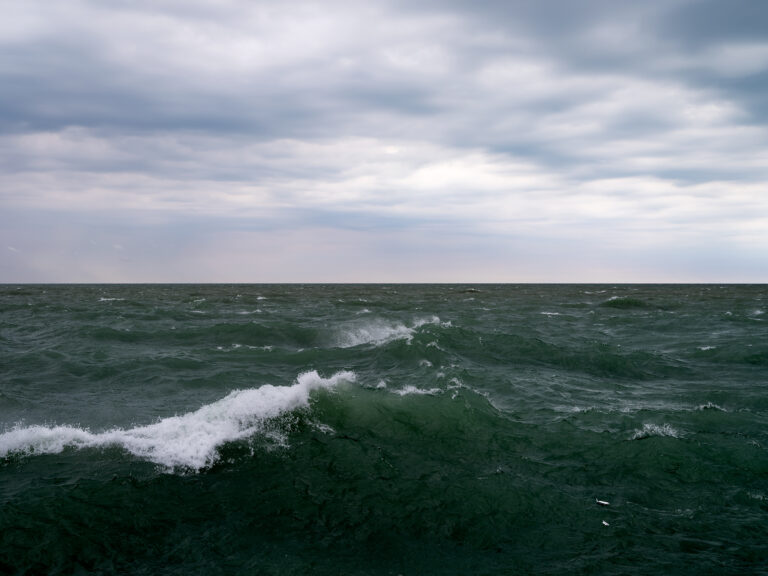 A curling wave in dark green lake water rolls to shore under cloudy skies in a photo from the Lake Series by Lincoln Schatz.