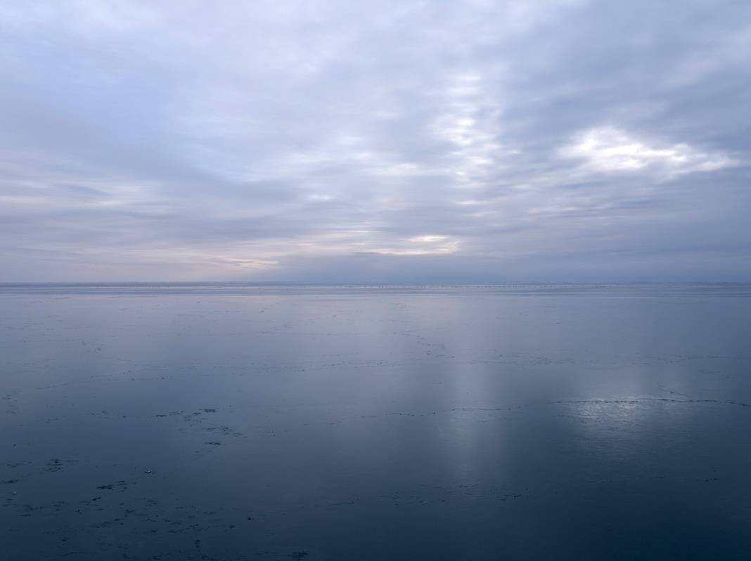The lake sits in absolute stillness as a result of a veneer of ice forming overnight. This thin barrier on the surface will disappear as soon as it has formed, its appearance following a series of storms. For now the lake reflects the gentle first light of day on the ice stretching away from my feet.