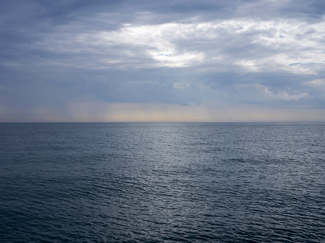 From the Lake Series, this is July 8th, 2024. The clouds are layering in the sky over Lake Michigan, creating soft and undulating patterns overhead. Rain falls out from shore, visible in a band above the horizon line. 