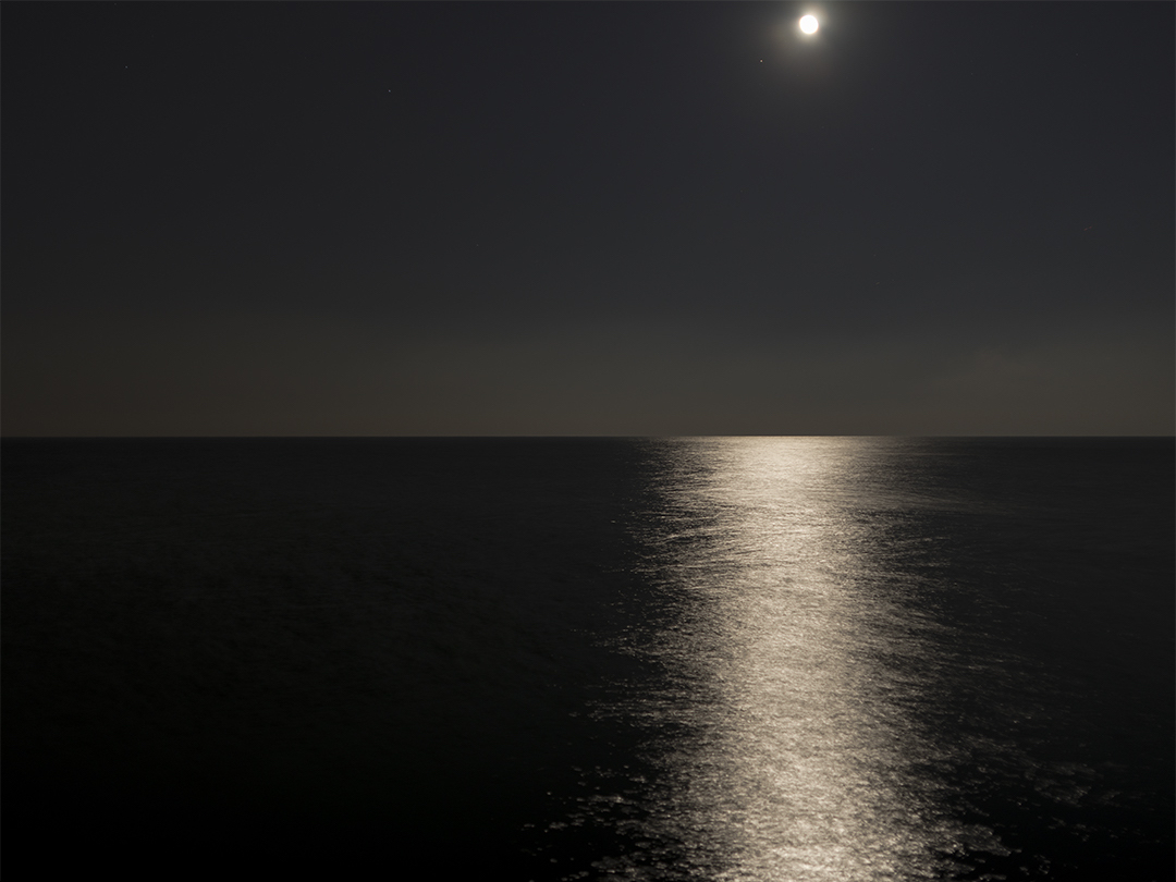 The moon is climbing into the sky over a calm Lake Michigan in a photograph from the Lake Series, December 12th, 2016. The water blurs in a beam of light reflecting off the surface of the lake.