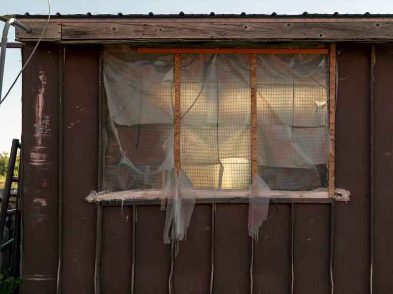 plastic sheeting in tattered layers fill the windows of a brown outbuilding